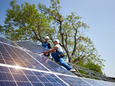 Two workers in protective helmet on steel platform installing heavy solar photo voltaic panel on green tree and blue sky background. Exterior solar panel system installation, dangerous job concept.